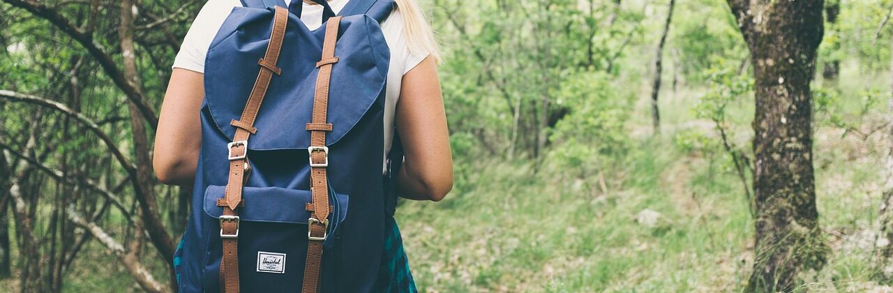 Blond woman with a backpack shown from behind hinking in the green woods.