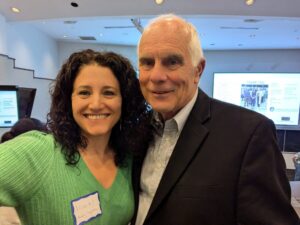 Galaxy Diagnostics CEO Nicole Bell, a white woman with black hair in a green sweater, and Cofounder and Chief Scientific Officer Dr. Ed Breitschwerdt, a white man with white hair in a black suit coat and a blue oxford shirt, pose together in a conference room. 